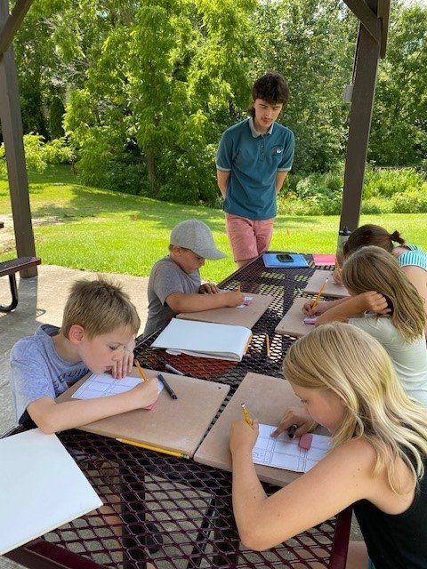Five children draw at an outdoor table while a young adult stands nearby, observing them.