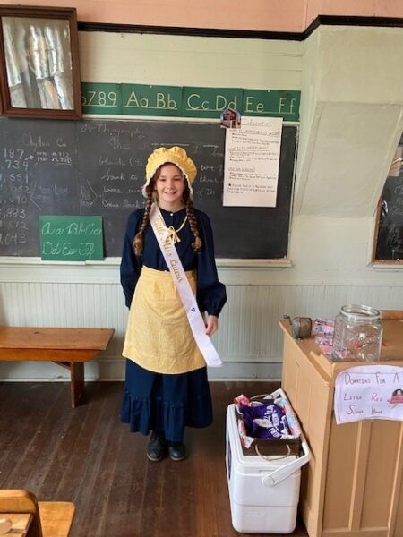 A girl in vintage clothes and a sash stands in an old-fashioned classroom with chalkboards and wooden desks.