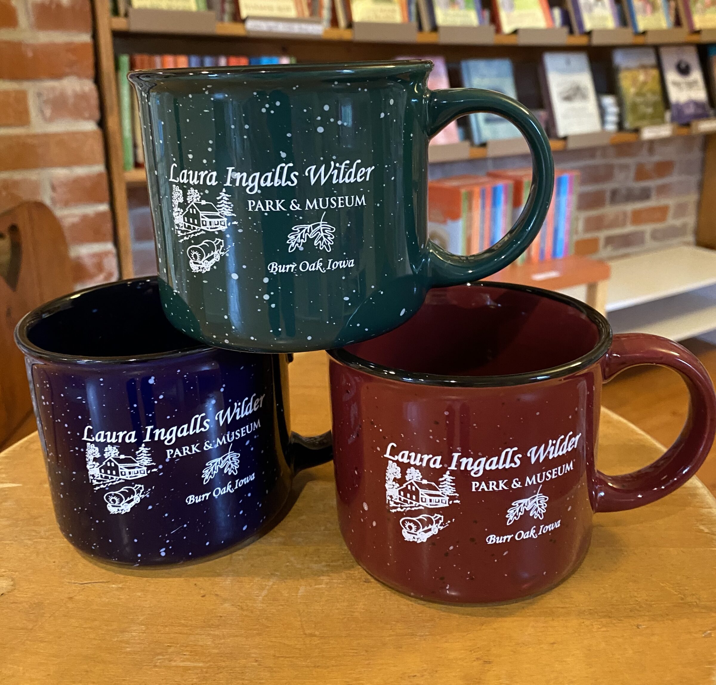Three Mugs from the Laura Ingalls Wilder Park & Museum are stacked on a table in front of bookshelves.