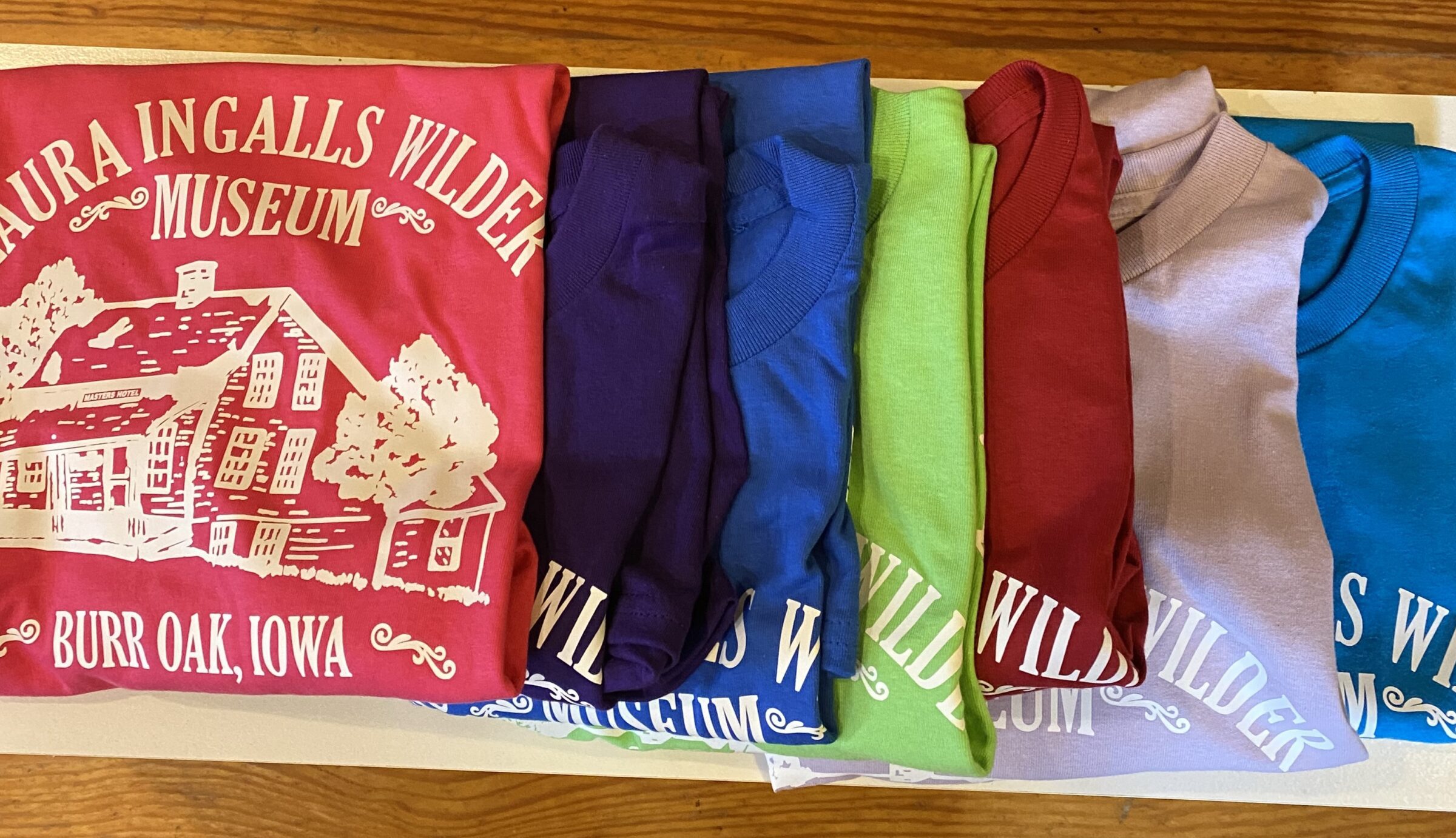 Colorful Adult T-shirts featuring “Laura Ingalls Wilder Museum” and a house graphic are displayed on a wooden surface.