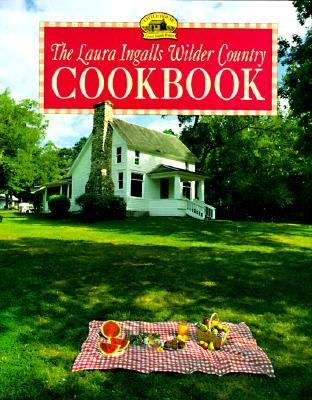 A white farmhouse with a stone chimney and a red checkered picnic blanket in front; cover of the Laura Ingalls Wilder Country Cookbook.