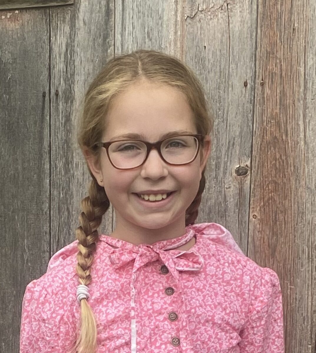 Smiling girl with glasses, braided hair, and a pink patterned dress stands in front of a wooden wall.