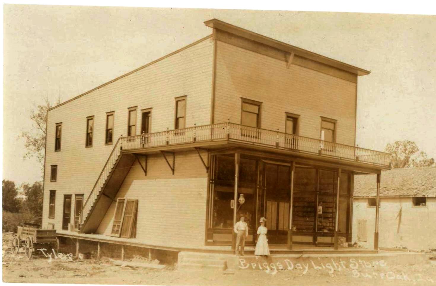 Sepia photo of a two-story wooden store with two people standing on the porch out front.