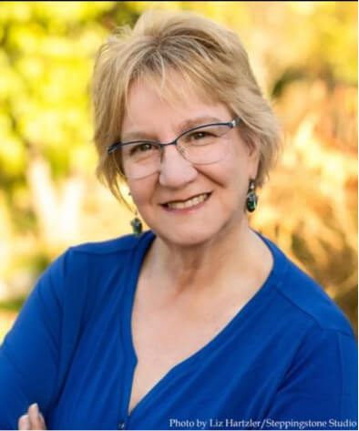 Smiling woman with short blond hair and glasses, wearing a blue top, outdoors with blurred greenery.