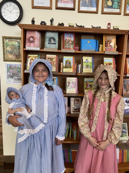 Two girls in pioneer-era dresses and bonnets stand in front of a bookshelf; one holds a matching doll.