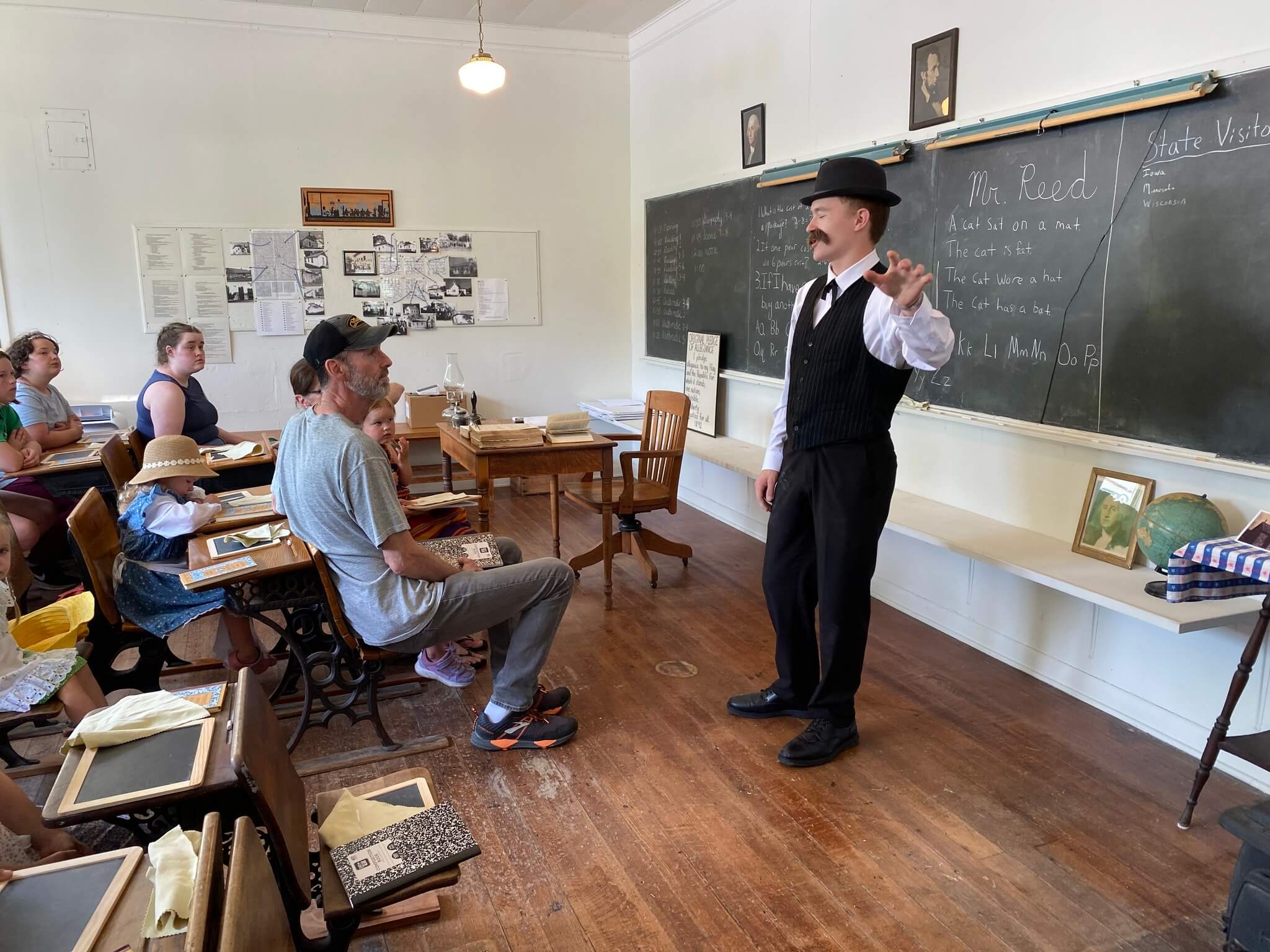 A man in vintage attire teaches children in an old-fashioned classroom with chalkboards and wooden desks.