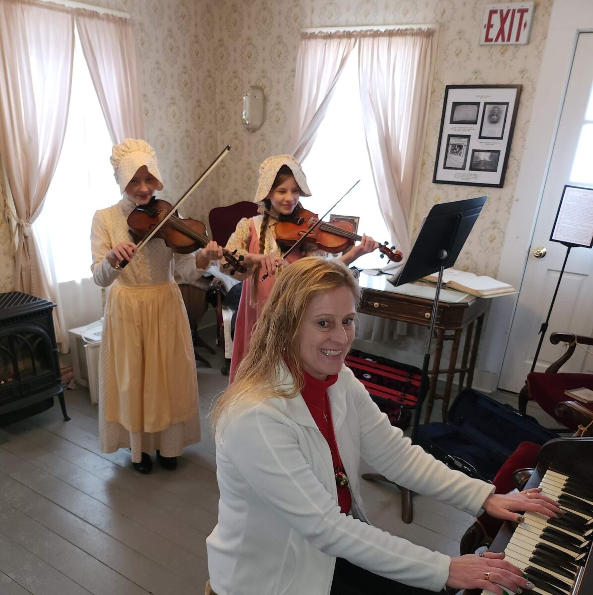 Two women in bonnets play violins, while a smiling woman in white plays piano in a vintage-style room.