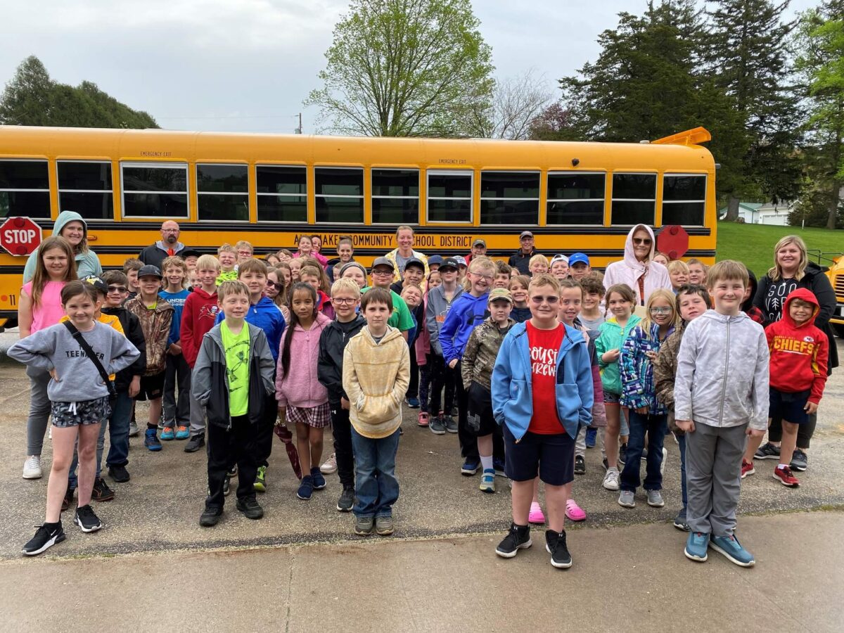 A large group of children and adults stand smiling in front of a yellow school bus.
