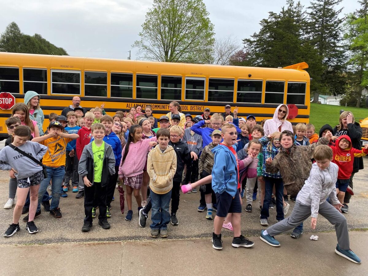 A large group of children pose playfully in front of a yellow school bus on a cloudy day.