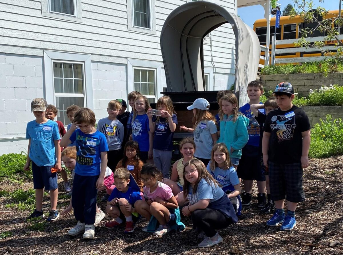 A group of children pose outside near a covered wagon, with a building and school bus in the background.