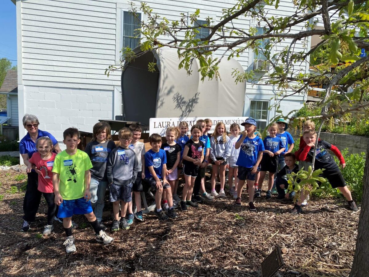 A group of children and an adult pose outside by a sign and garden on a sunny day.