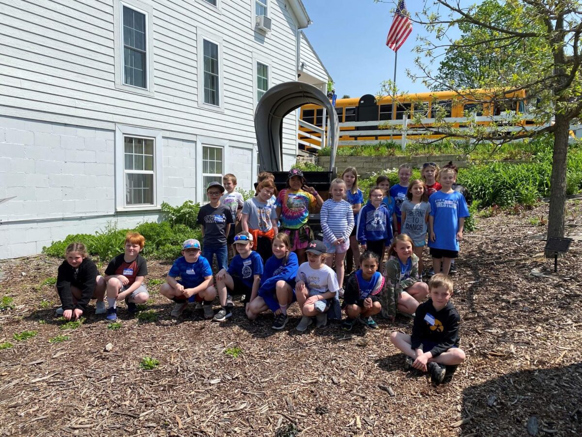 A group of children pose outside a building near a yellow school bus on a sunny day.
