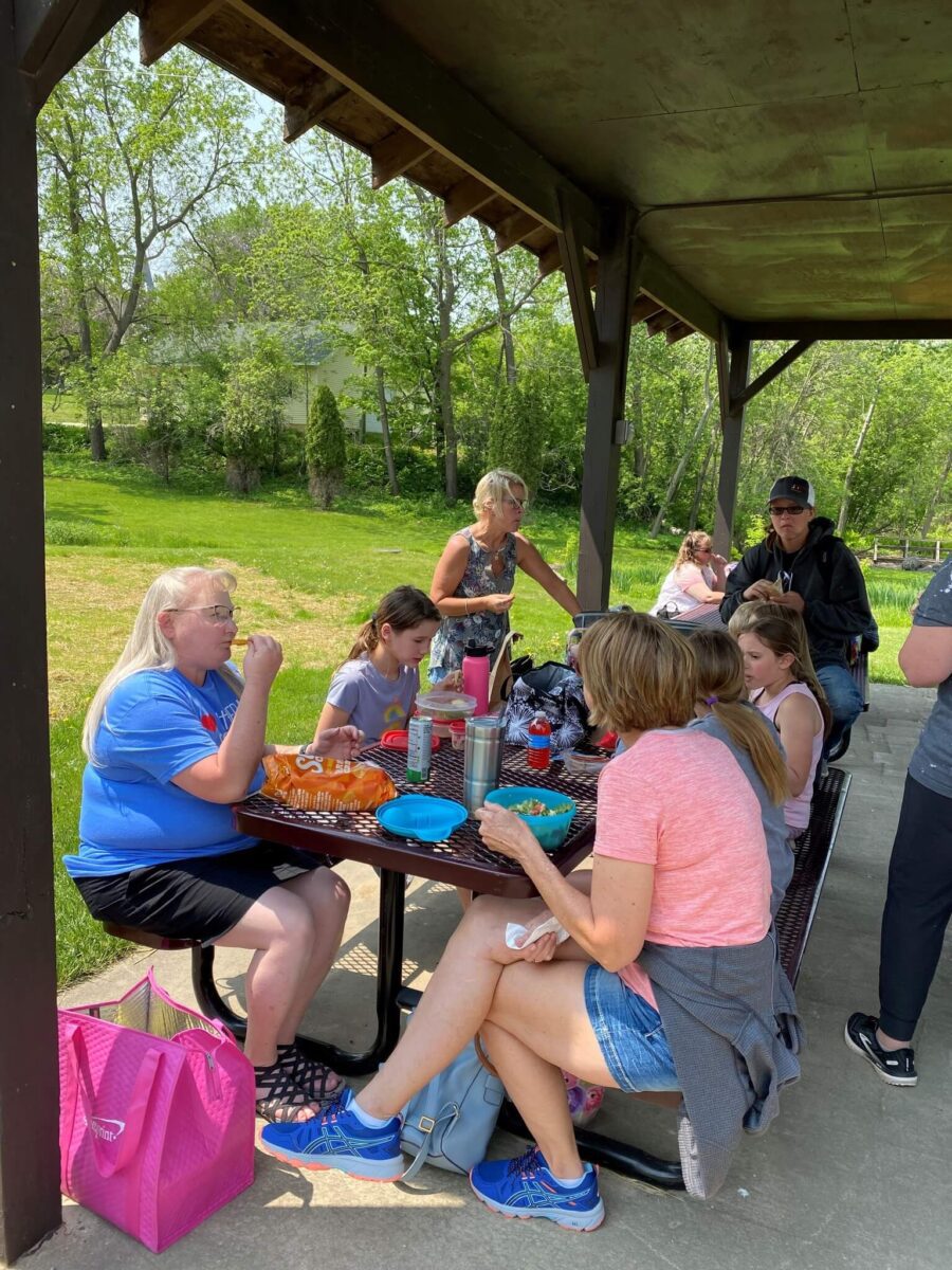 A group of people sit at a picnic table under a shelter, sharing food and drinks on a sunny day.