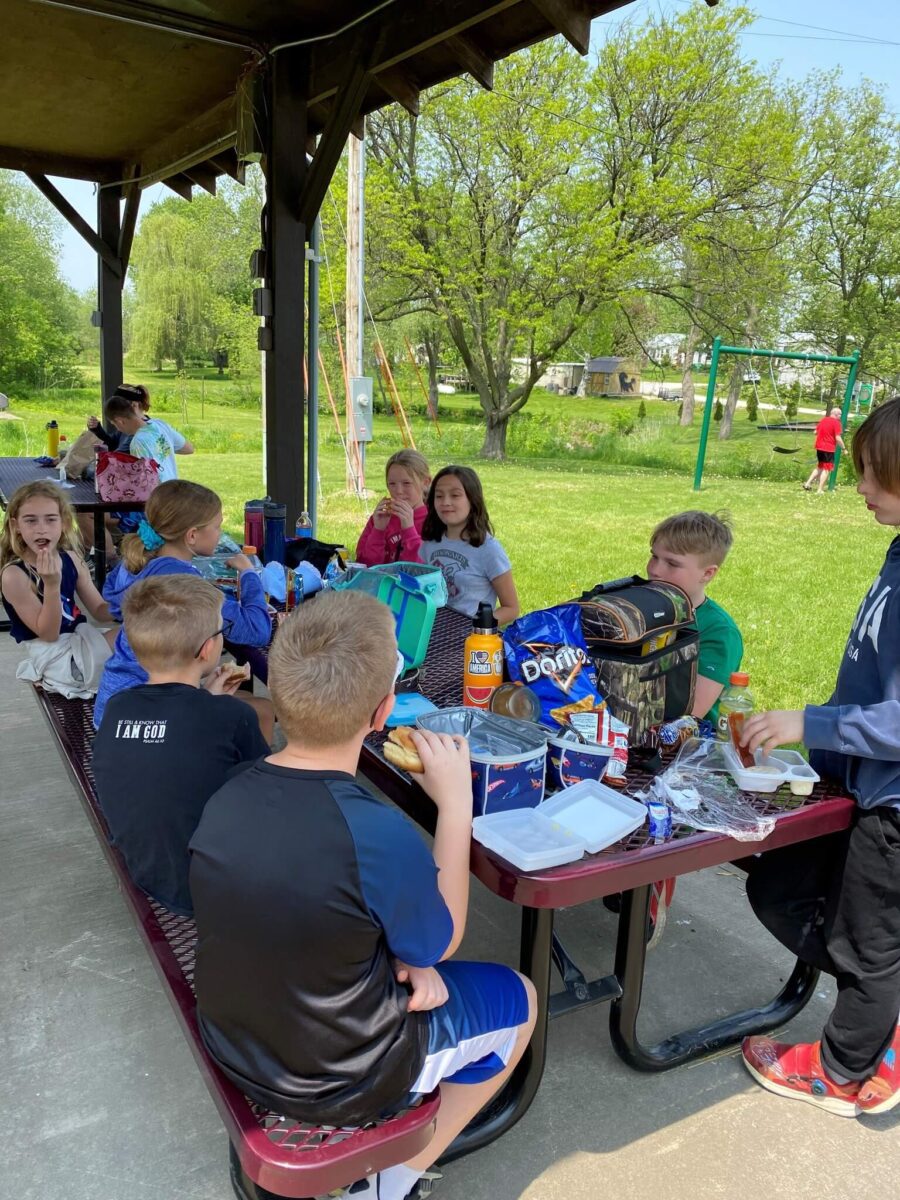 A group of kids eat lunch together at a picnic table under a pavilion in a park on a sunny day.