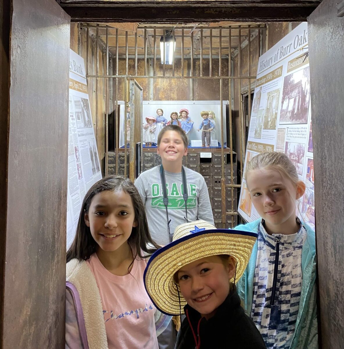 Four smiling children stand inside an old jail cell, posing in front of historical display boards.