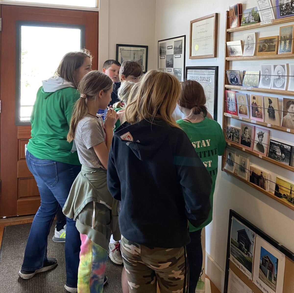 A group of kids in casual clothes look at photos and documents on a wall inside a building.