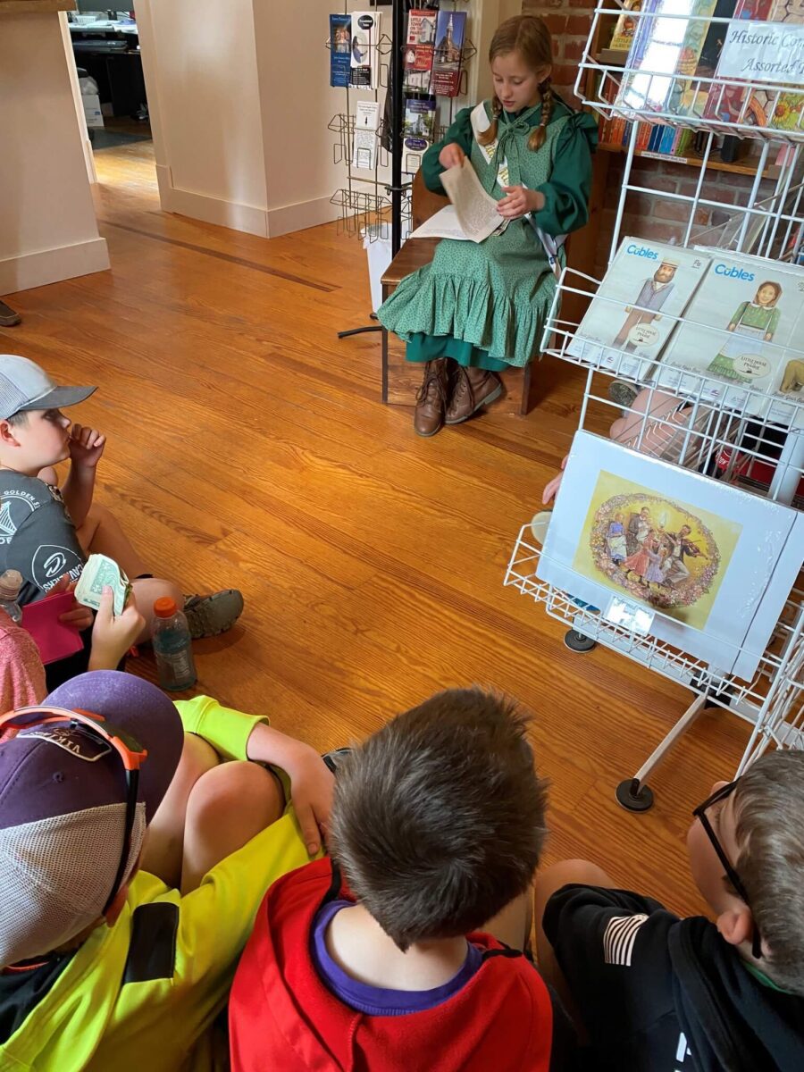 A woman in a green dress reads to a group of children sitting on the floor in front of her.
