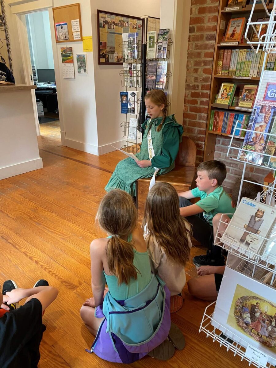 A girl in a green dress reads aloud to children sitting on the floor in a library.