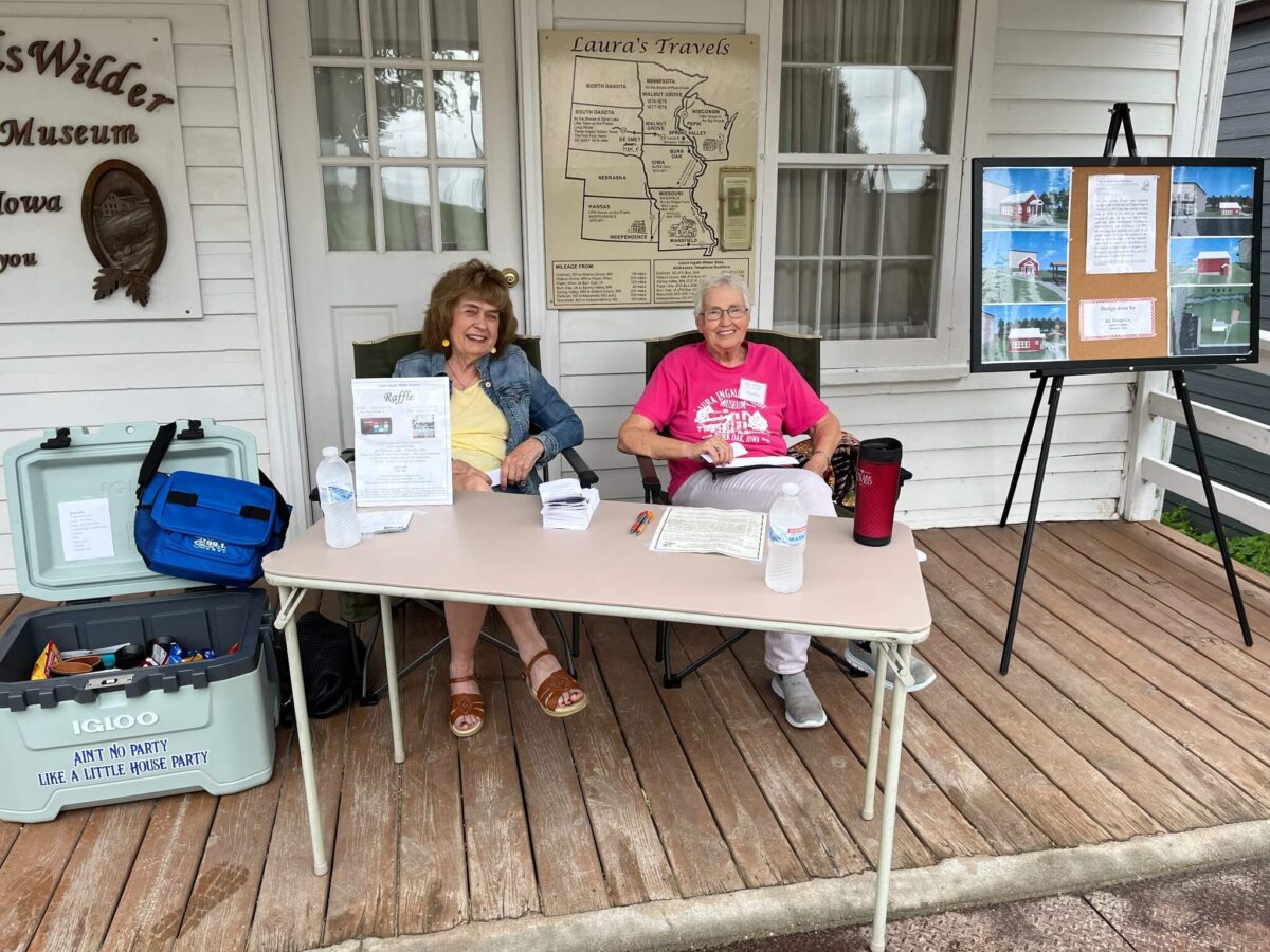 Two smiling women sit at a table with papers outside a museum on a wooden porch, welcoming visitors.