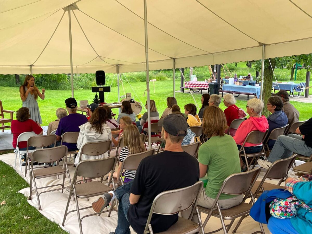 A woman speaks to an audience seated under a white tent at an outdoor event in a park.