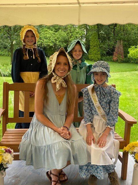 Four girls in pioneer-style dresses and bonnets pose on a bench outdoors, smiling under a canopy.