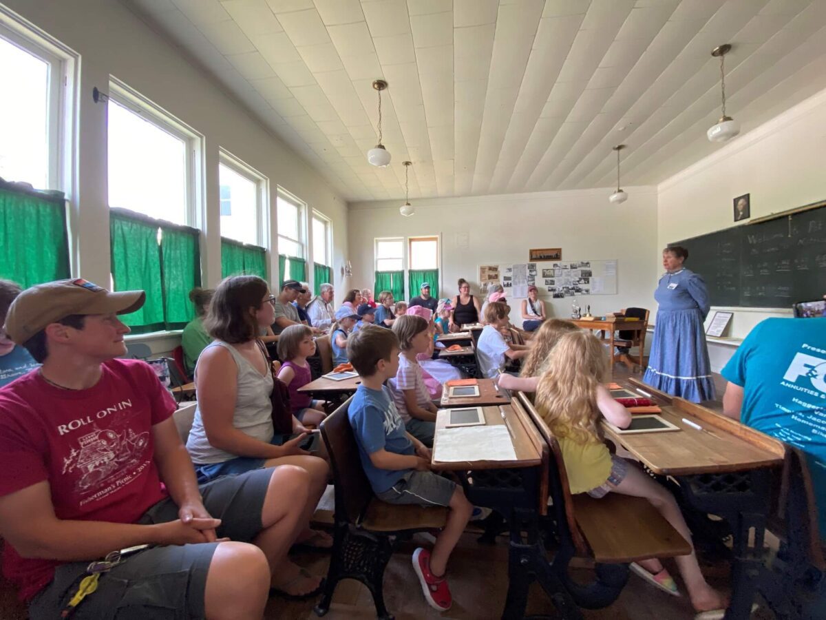 A woman in period dress teaches children and adults in a historic classroom with wooden desks and green curtains.
