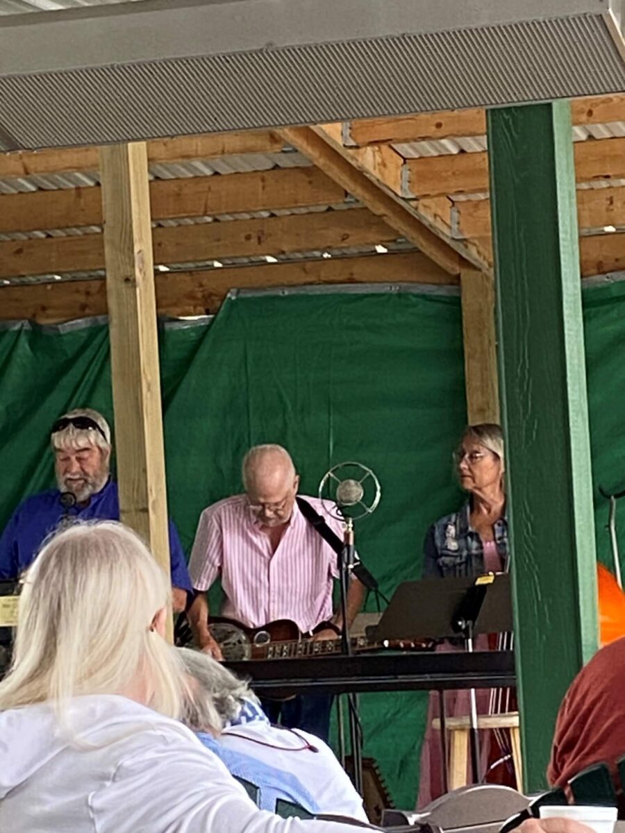 Three musicians perform on a wooden stage with a green backdrop, watched by an audience.