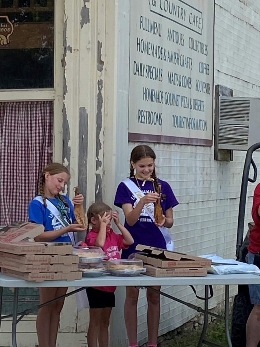 Three girls with sashes eating pizza stand next to a table with pizza boxes outside a vintage-style café.