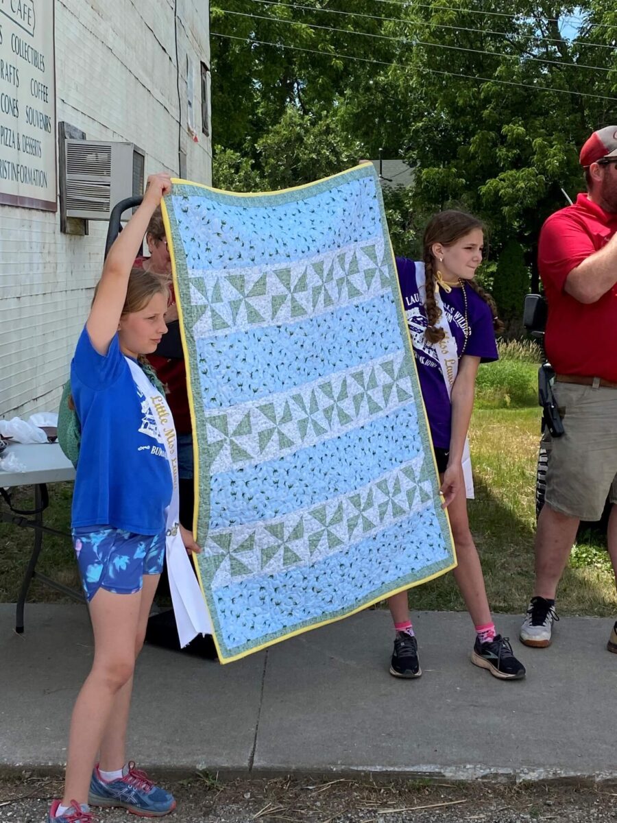 Two girls hold up a blue and green quilt outside, while a man in red stands nearby speaking.