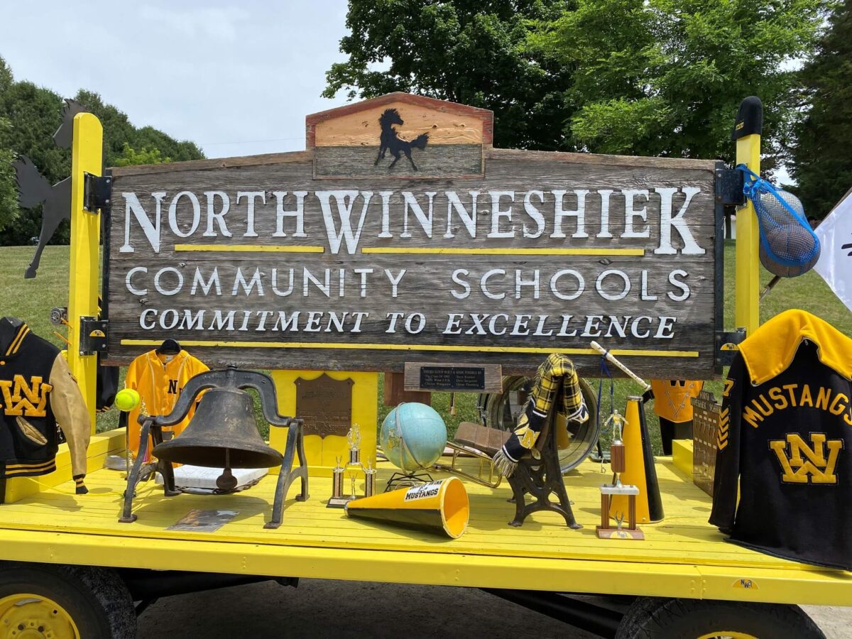 North Winneshiek Community Schools sign decorated with school memorabilia on a yellow cart outdoors.