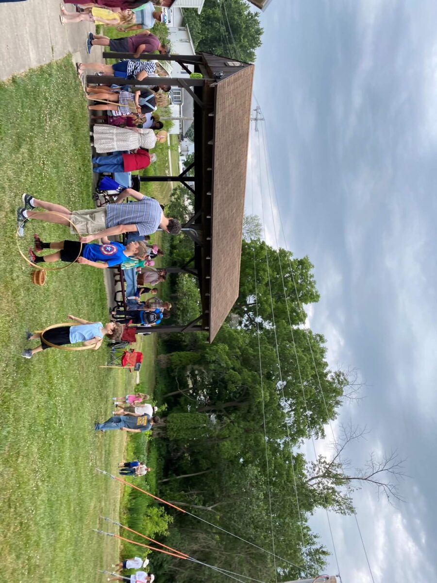 People gather near a pavilion at a park, with children playing on the grass under a cloudy sky.