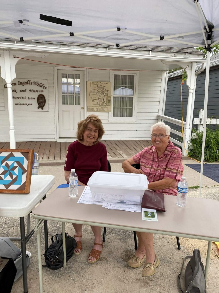 Two older women smile at a table with papers and water bottles outside a white building under a canopy.