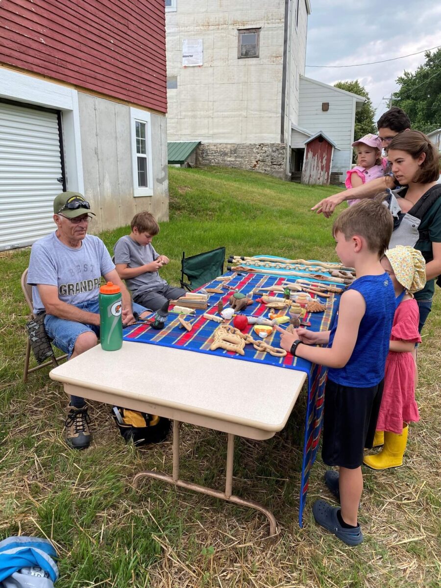 Children and adults browse handmade wooden toys on a colorful table outdoors, with an older man seated nearby.