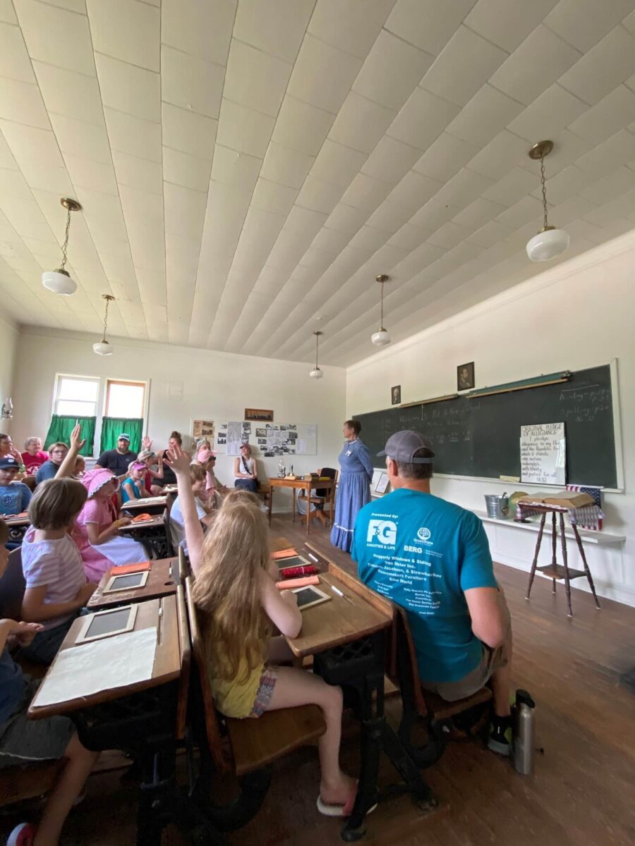 Children and adults sit at desks in a vintage classroom as a woman at the front leads a lesson.