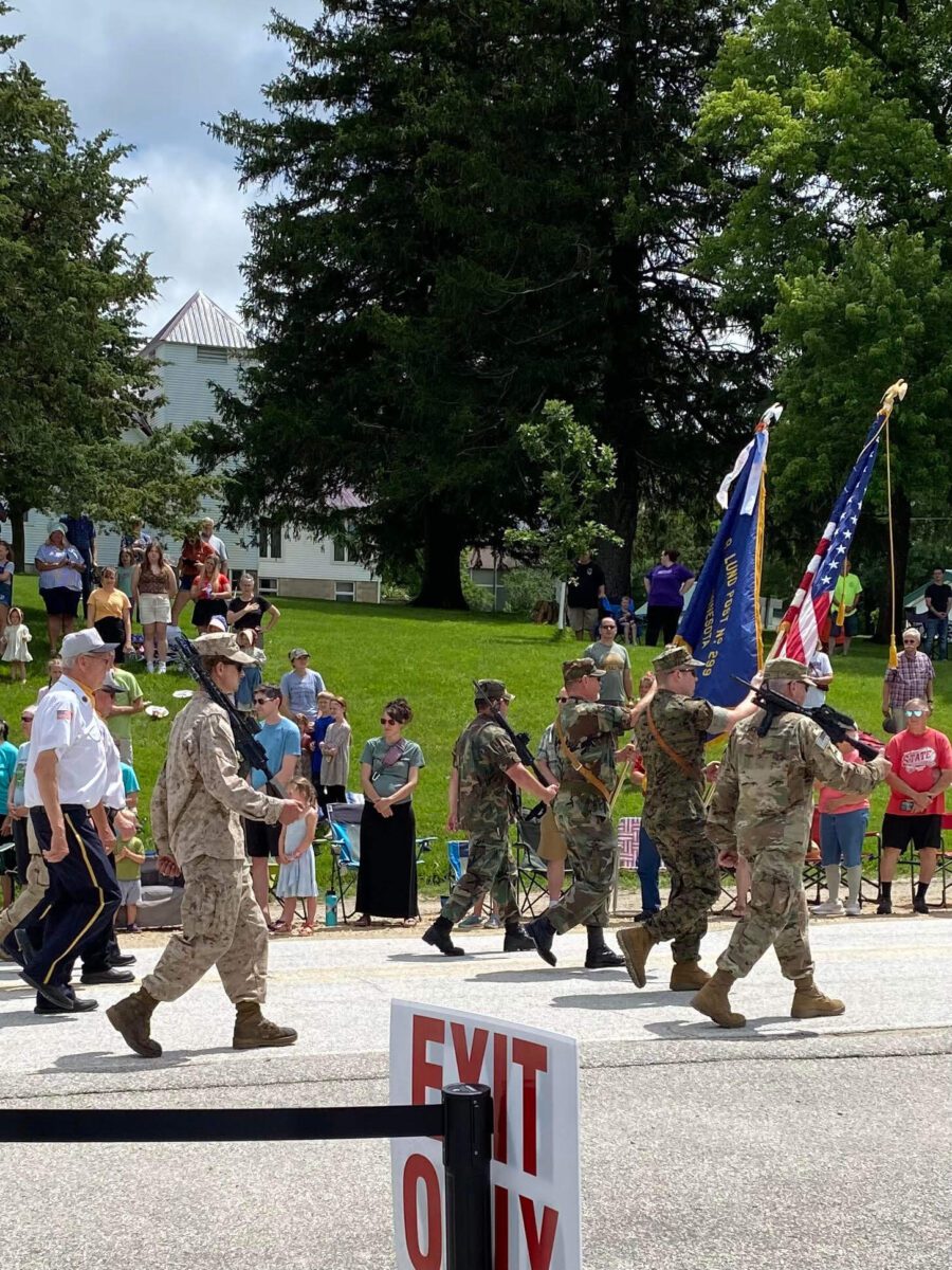 Military veterans in uniform march in a parade, carrying flags, with spectators watching from the roadside.
