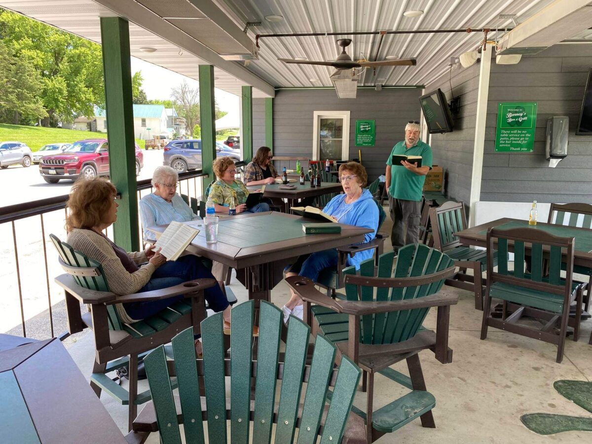 A group of people sits outside at tables reading, with one person standing and speaking to them.