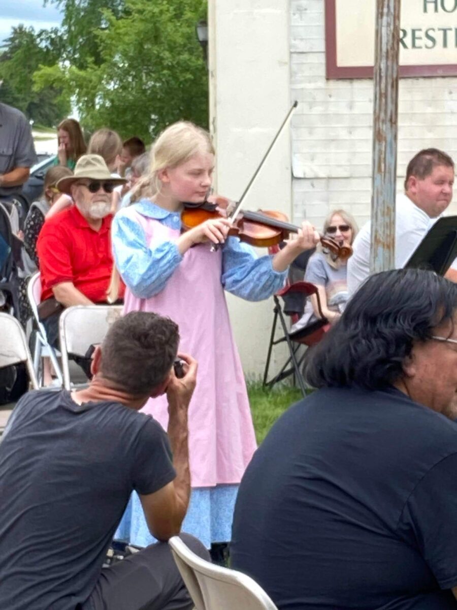 A young girl in a pink dress plays violin outdoors as people watch and a photographer kneels in front.