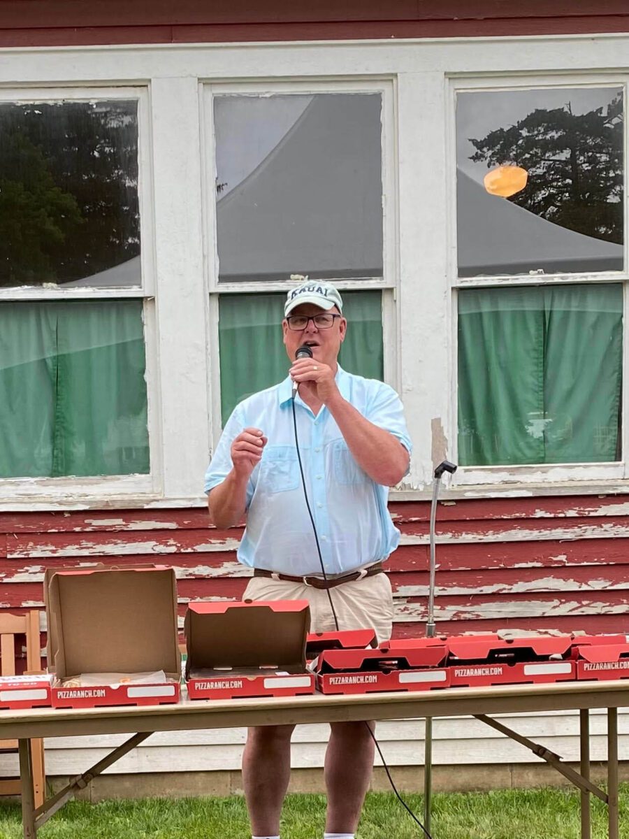 Man in a cap speaks into a microphone outside, standing behind a table with open pizza boxes.