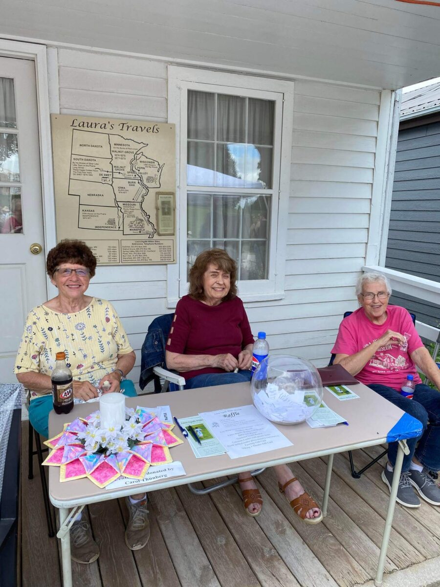 Three smiling women sit at a table with raffle tickets and drinks on a porch in front of a map display.