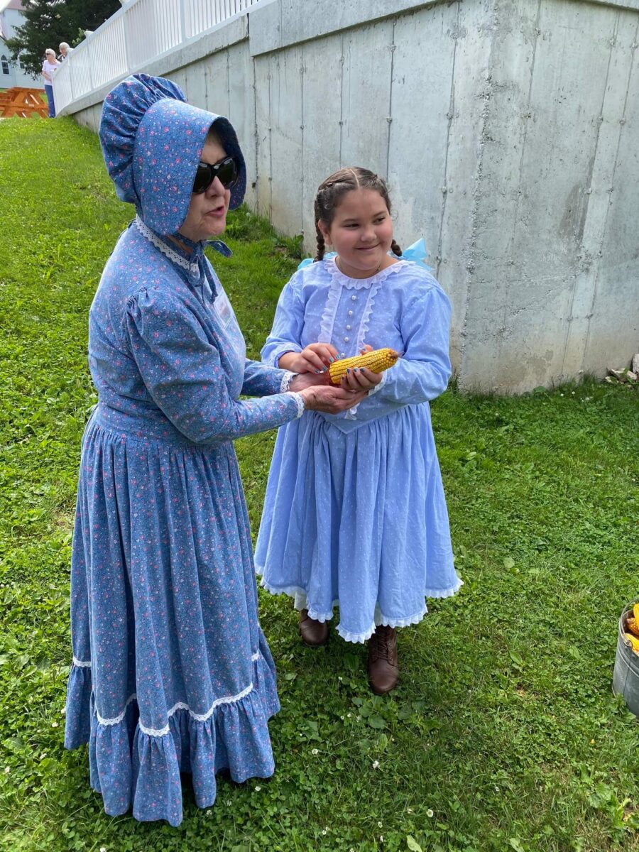 Two people in blue pioneer dresses stand on grass; one hands an ear of corn to the other, both smiling.