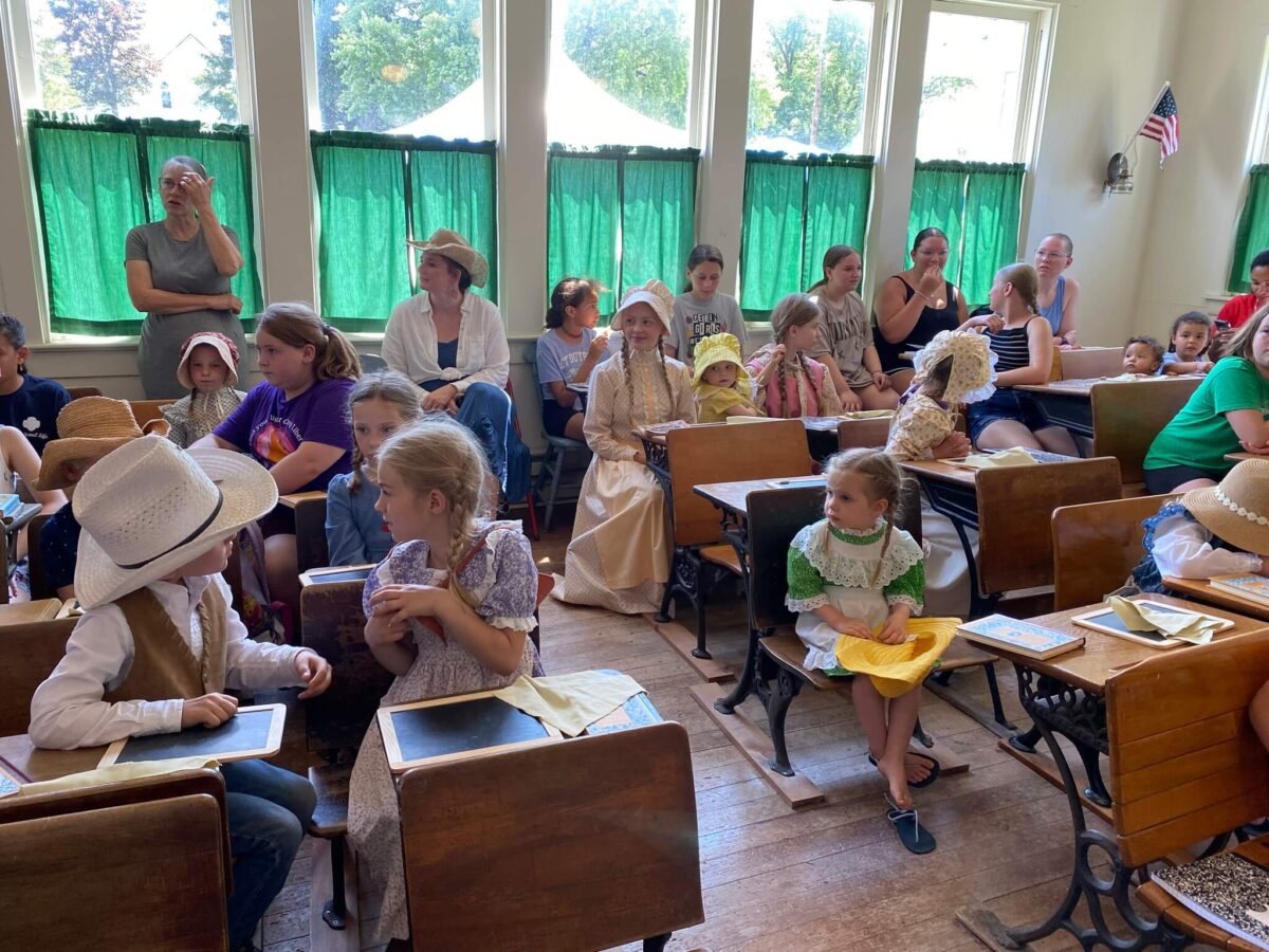 Children and adults in period costumes sit in an old-fashioned classroom with green curtains and wooden desks.