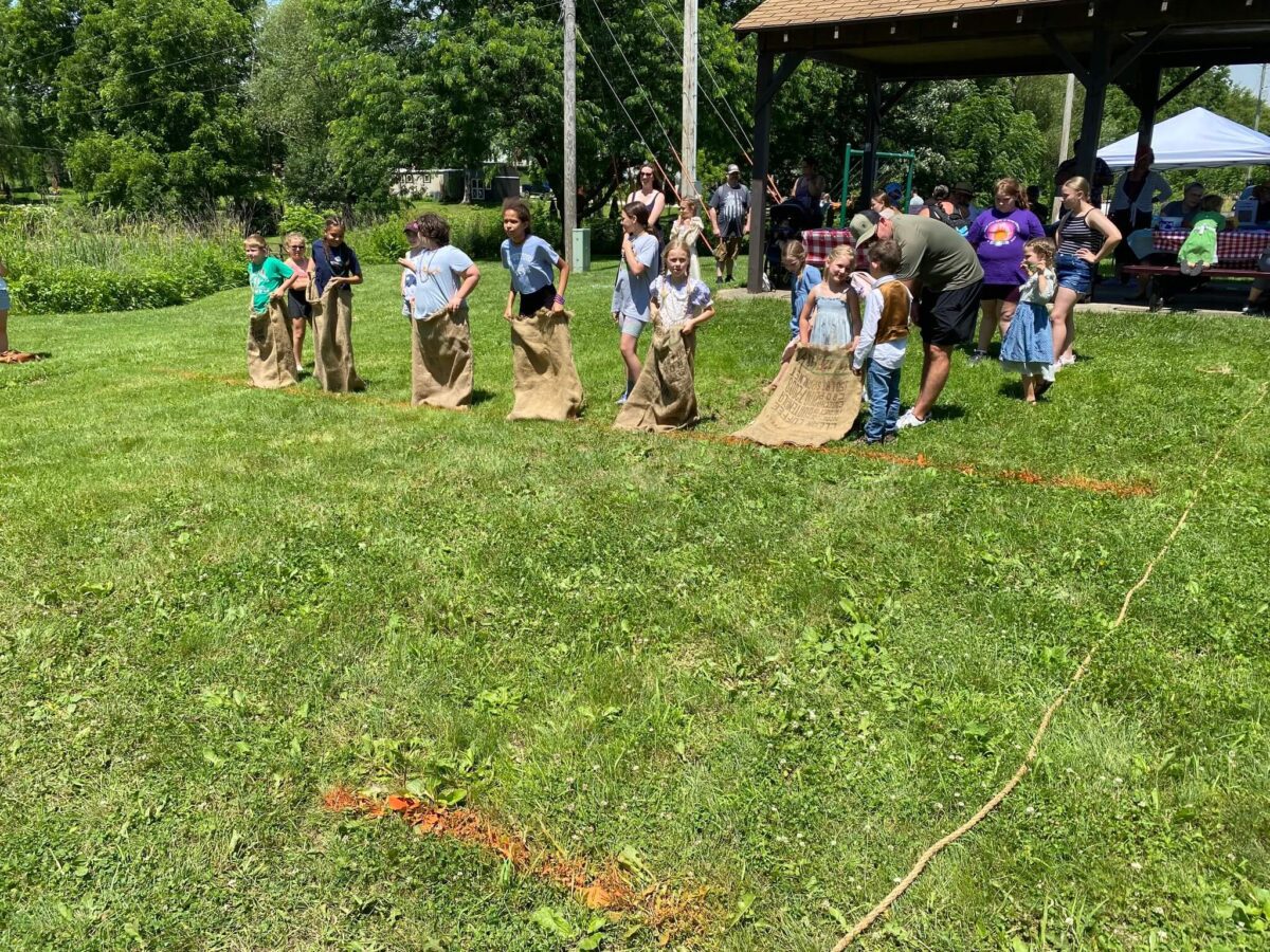 Children line up in burlap sacks for a sack race on a sunny grassy field at an outdoor event.