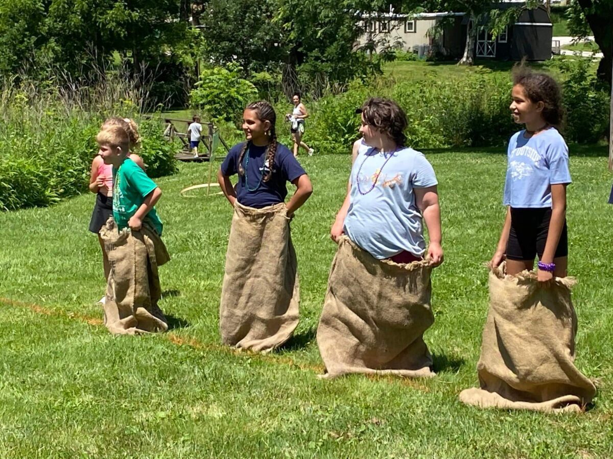 Four children stand in burlap sacks on a grassy field, ready for a sack race on a sunny day.