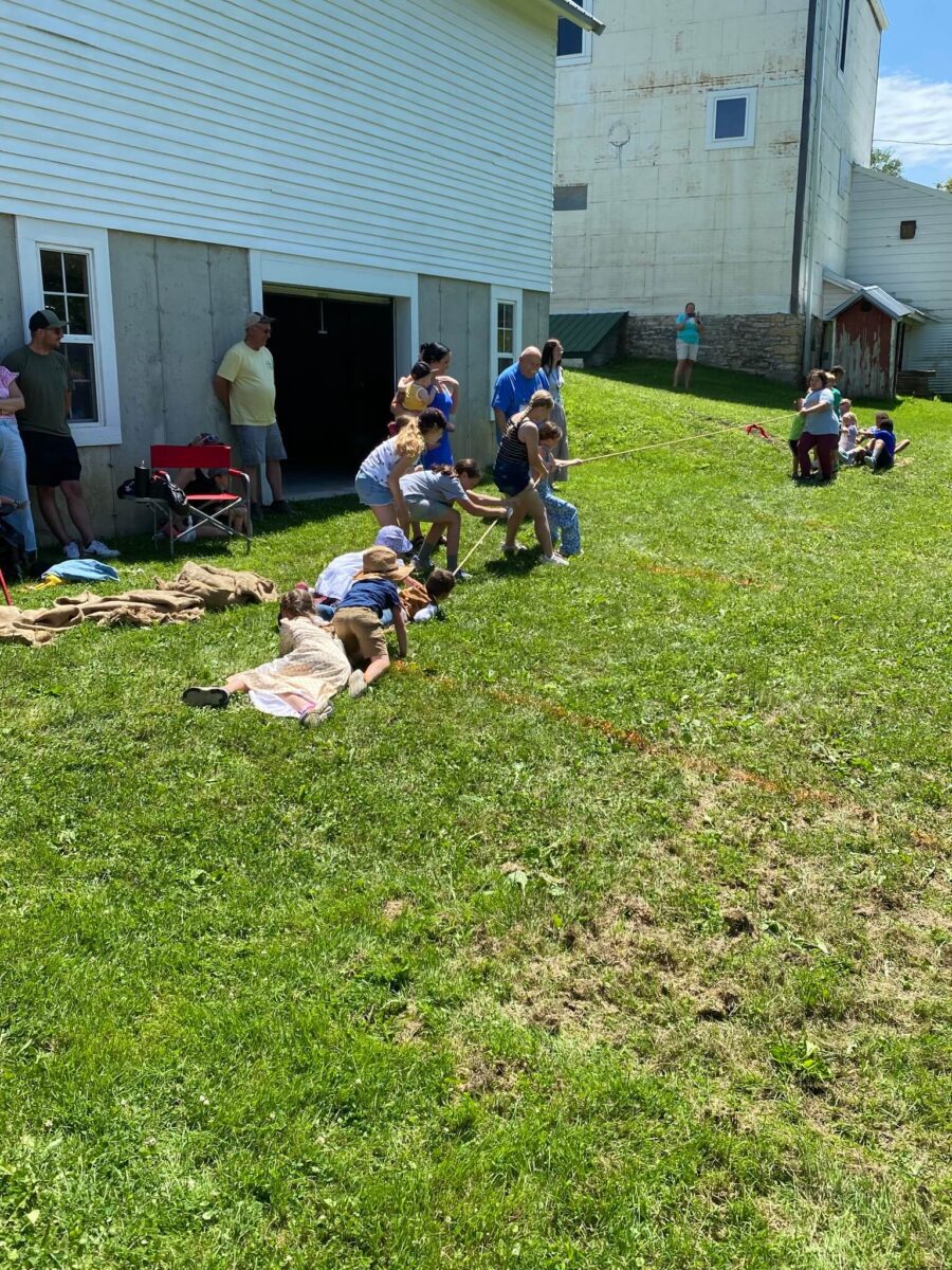 Children and adults play tug-of-war on a grassy lawn near a white building on a sunny day.