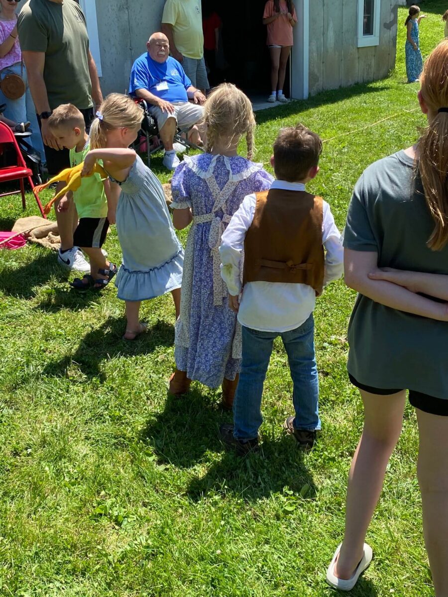 Children stand in line outside on grass, some wearing costumes, while adults watch nearby in the sunlight.