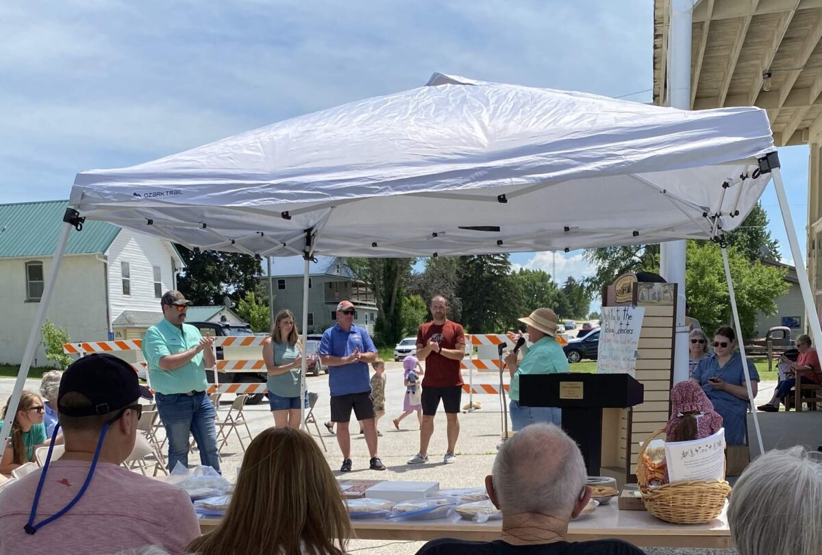 People gather under a white tent at an outdoor event with a speaker at a podium and baked goods on a table.