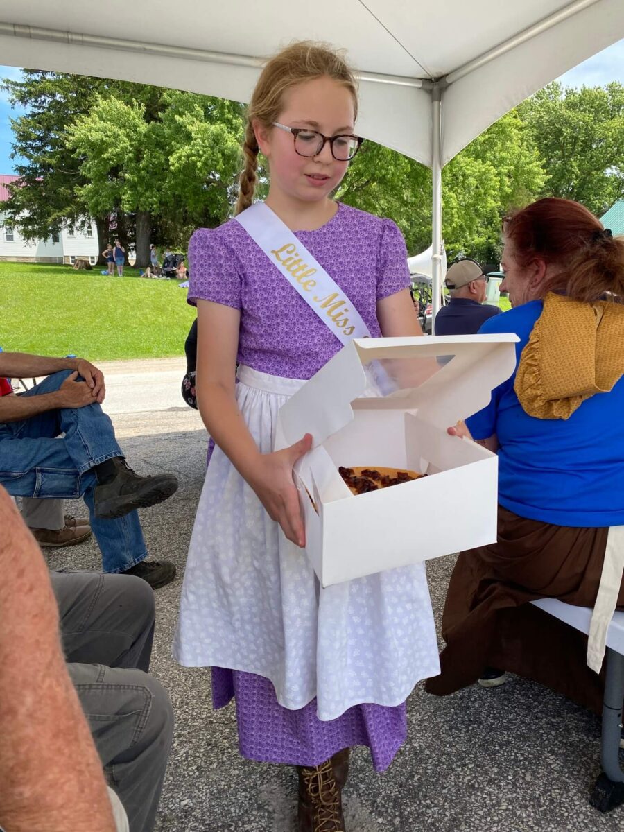 A girl in a sash and purple dress holds an open bakery box with a pie at an outdoor event.