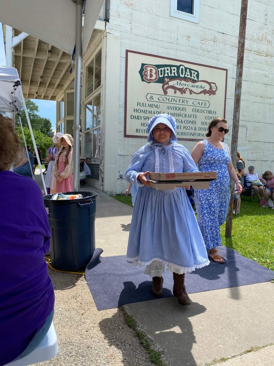 Girl in pioneer dress carries pizza boxes at outdoor event near Burr Oak Mercantile sign on a sunny day.