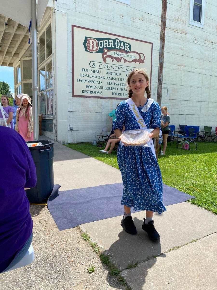 Young girl in a blue dress and sash holds a pie outside Burr Oak Mercantile, people and sign in background.