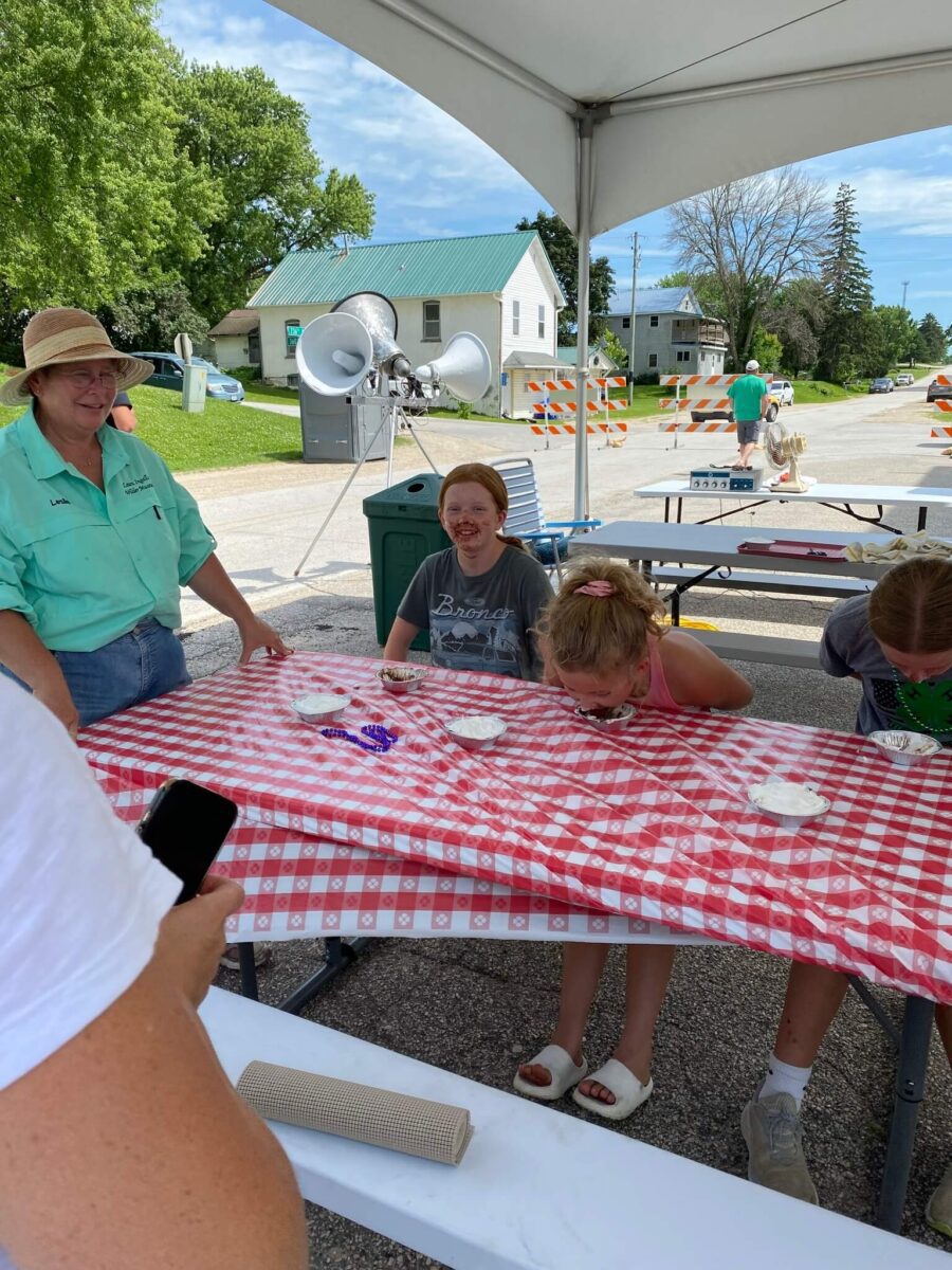 Children compete in a pie-eating contest at an outdoor event under a tent, with people watching and smiling.